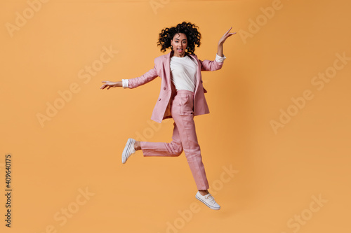 Enthusiastic mixed race girl jumping on yellow background. Full length view of gorgeous young woman in pink suit.