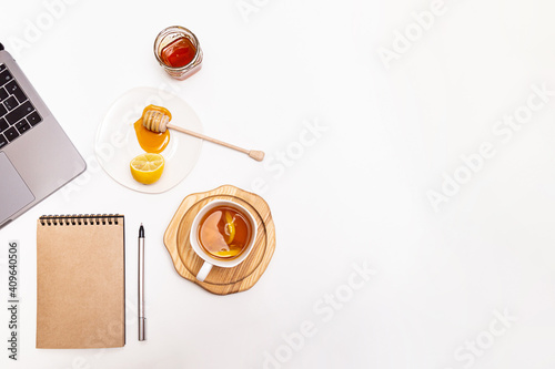 Cozy home work place. A cup of tea with honey and lemon, a laptop and a notebook with a pen on a white table with a place for text.