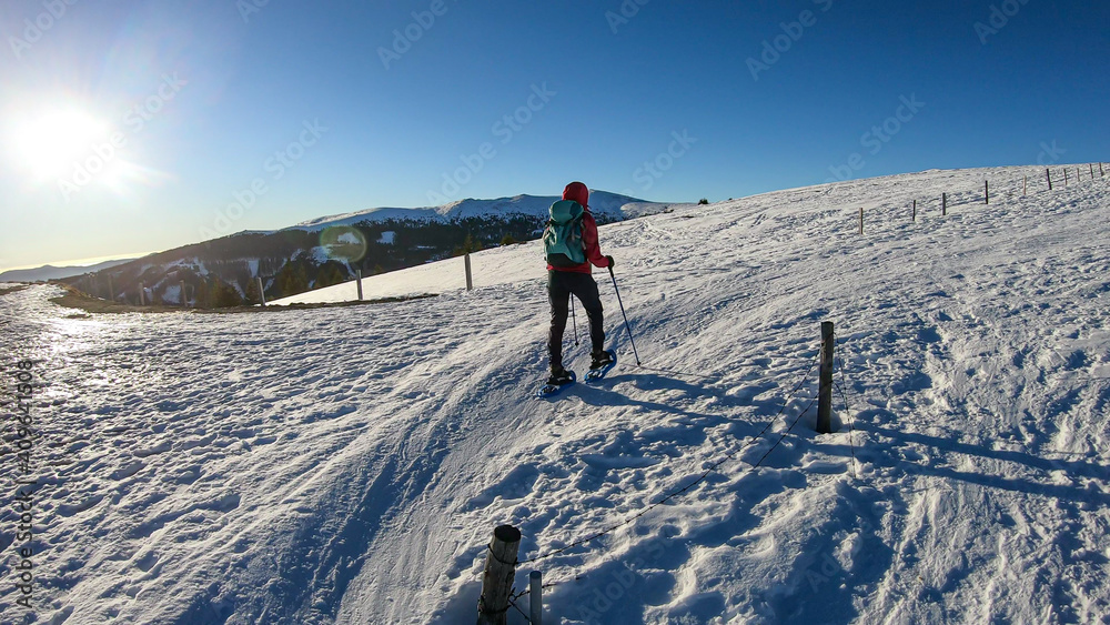 A woman walking on the snow shoes up to Amerinkogel's peak in Austrian Alps through a vast pasture covered with powdered snow. Many mountain chains in the back. Winter outdoor activity. Fun and chill