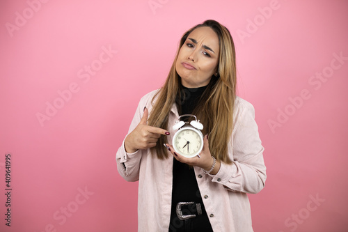 Young beautiful blonde woman with long hair standing over pink background screaming and scared, pointing the clock, because it's late