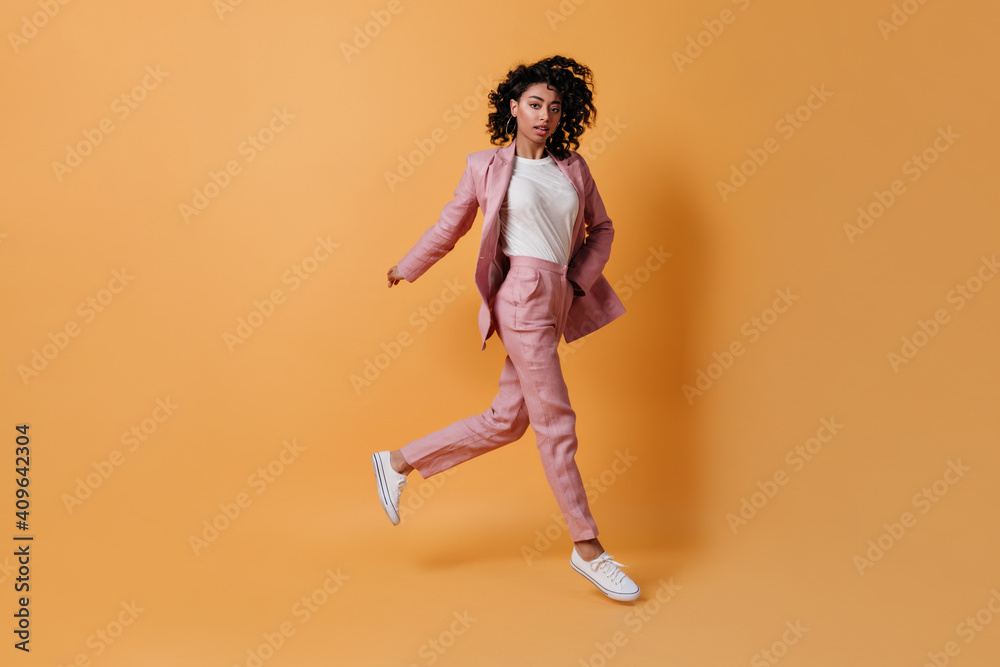 Full length view of running curly woman. Adorable girl in pink suit jumping on yellow background.