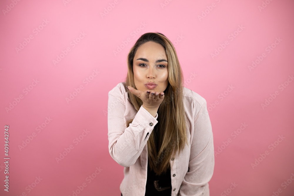 Young beautiful blonde woman with long hair standing over pink background looking at the camera blowing a kiss with hand on air being lovely and sexy. Love expression. Young beautiful blonde woman with long hair standing over pink background looking at the camera blowing a kiss with hand on air being lovely and sexy. Love expression.