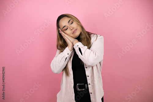 Young beautiful blonde woman with long hair standing over pink background sleeping tired dreaming and posing with hands together while smiling with closed eyes.