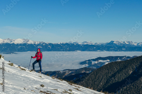 A woman wearing pink jacket and snow shoes hiking up to Amerinkogel's peak in Austrian Alps. Fresh powder snow. Many mountain chains in the back, valley shrouded in fog. Winter outdoor activity. Fun photo