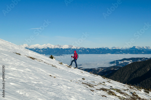 A woman wearing pink jacket and snow shoes hiking up to Amerinkogel's peak in Austrian Alps. Fresh powder snow. Many mountain chains in the back, valley shrouded in fog. Winter outdoor activity. Fun