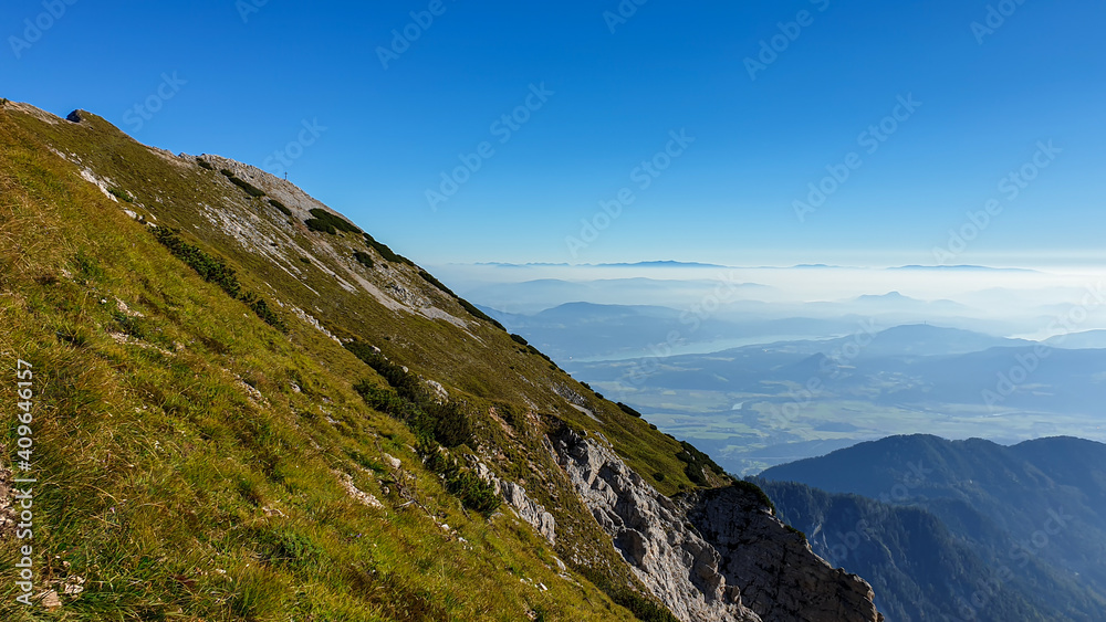 Panoramic view on the haze shrouded valley from the way to Mittagskogel in Austrian Alps. Clear and sunny day. Endless mountain chains. Outdoor activity. Barren top of the mountains, lush lower parts