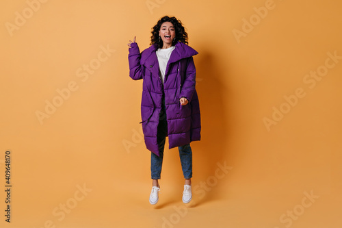 Smiling mixed race girl gesturing on yellow background. Emotional woman in purple down jacket jumping and looking at camera.
