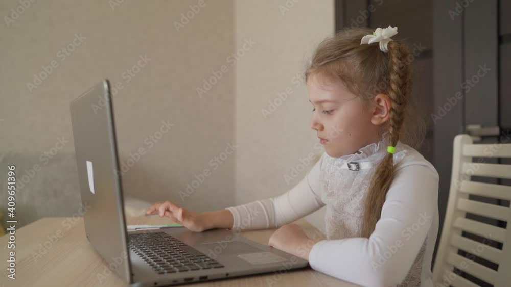 A little girl is sitting at home at a laptop. Medium shot of a cheerful little girl sitting at a table at home. A little girl is sitting at home at a laptop. Medium shot of a cheerful little girl sitting at a table at home.