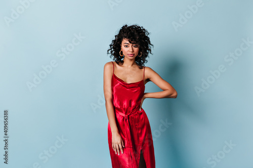 Charming curly young woman in red dress looking at camera. Studio shot of confident mixed race girl isolated on blue background.