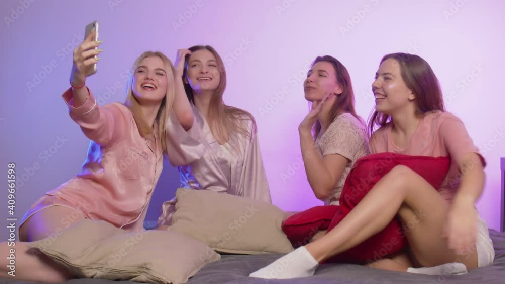 Four happy pretty young women in pajamas sits on bed and takes selfies on phone at bachelorette party Four happy pretty young women in pajamas sits on bed and takes selfies on phone at bachelorette party