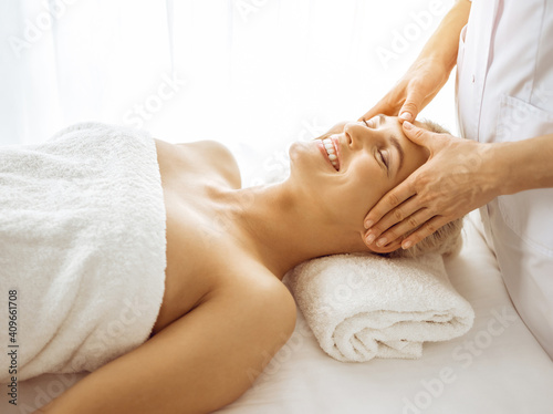 Beautiful blonde woman enjoying facial massage with closed eyes in sunny spa center. Relaxing treatment in medicine and beauty concepts