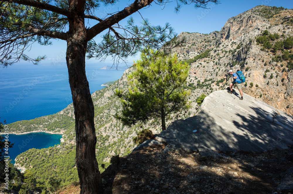 Hiking on Lycian way. Man with backpack looking down from steep rock high above Mediterranean sea, Trekking in Turkey on Lycian Way trail, outdoor activity Hiking on Lycian way. Man with backpack looking down from steep rock high above Mediterranean sea, Trekking in Turkey on Lycian Way trail, outdoor activity