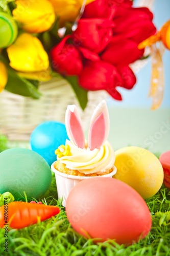 Easter funny bunny cupcake with colorful eggs. Easter celebration festive table. Basket of flowers tulips on the background.