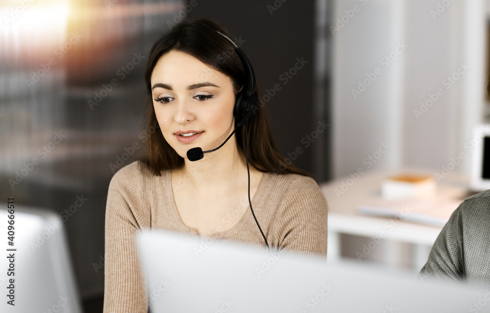 Young friendly girl in headsets is talking to a firm's client, while sitting at the desk in sunny office. Call center operators at work