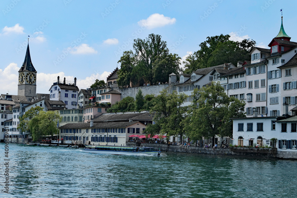 Zurich, Switzerland, June 30, 2018:  A tour boat cruises along the Limmat River the clock tower of the Church of St. Peter and the spire of the Augustinian Church in the background.