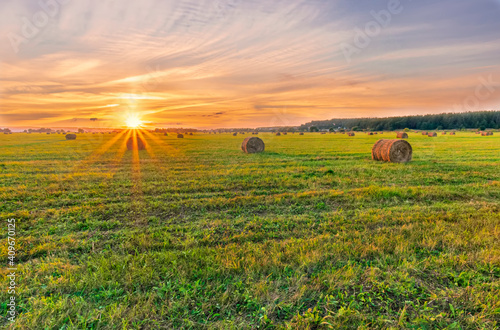 Scenic view at beautiful sunset in a green shiny field in willage farm with hay stacks, cloudy sky, golden sun rays, anazing summer valley evening landscape