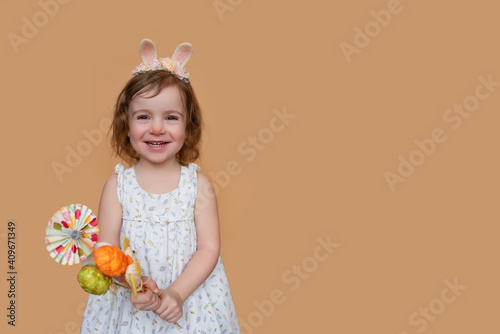 Emotional  positive portrait of little girl with bunny ears on head holding an Easter bouquet of eggs  chickens. Isolated light beige background place for text cards copy space. Festive child costume