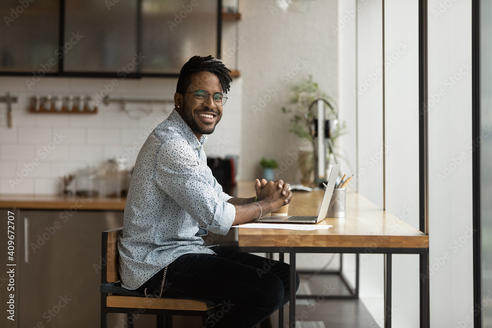 Portrait of happy afro american young male freelancer working by laptop at cozy home office at kitchen. Smiling positive black millennial man hipster looking at camera distracted from computer screen