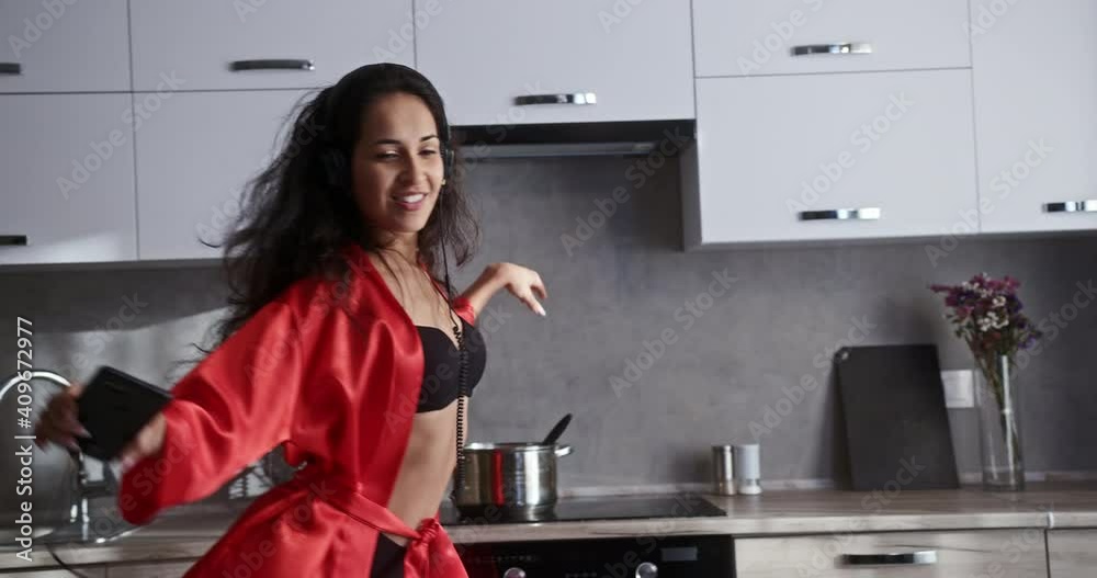 Young beautiful dancer girl with curly hair dances at home during quarantine to music in headphones in a bright kitchen in a beautiful red robe Young beautiful dancer girl with curly hair dances at home during quarantine to music in headphones in a bright kitchen in a beautiful red robe
