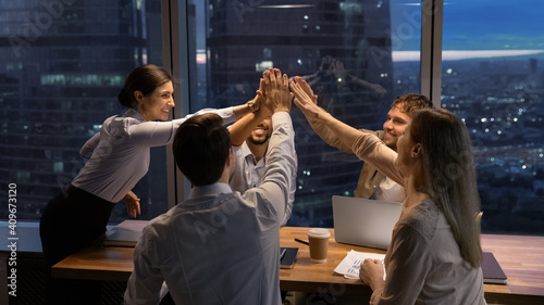 Motivated multinational team raise high fives on briefing after finding problem solution as successful brainstorm result. Happy workers unite hands above conference desk celebrate common achievement photo