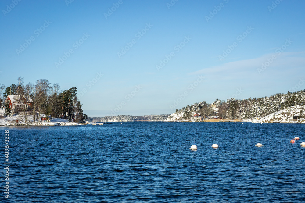 Winter landscape of the Swedish coast. The shore of Baltic sea overgrown with pines and firs covered with snow. Traditional  red wooden house surrounded by forest. Scandinavia in the winter season.