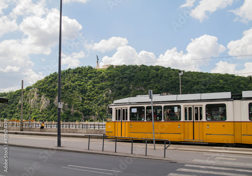 Streets of Budapest. Yellow streetcar against the background of Gellert Hill. Green trees and blue sky. Sunny day in Hungary.