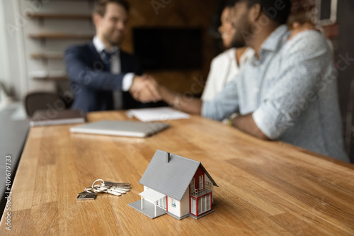 Focus on close up keys bunch and cottage house toy model on wooden table. Smiling young man broker realtor real estate agent shake hands of happy black couple clients homeowners on blurred background photo