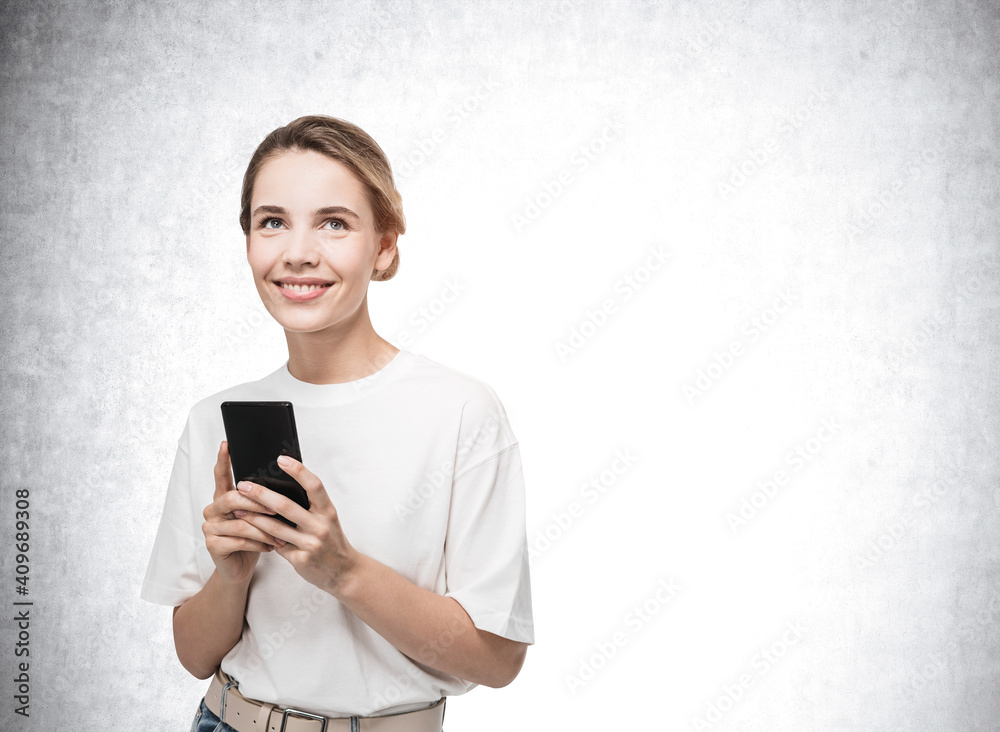 Attractive young woman on concrete background holding smartphone smiling and dreaming about pleasant things Attractive young woman on concrete background holding smartphone smiling and dreaming about pleasant things