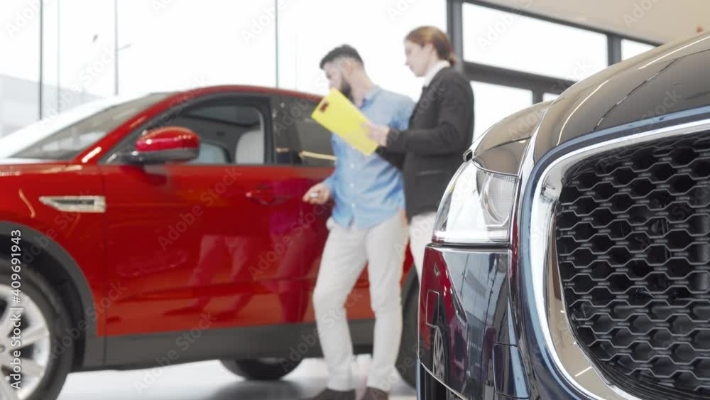 Salesman and male customer examining automobile at car dealership. Selective focus on a car on foreground, car dealer talking to a client on background. 4k footage Salesman and male customer examining automobile at car dealership. Selective focus on a car on foreground, car dealer talking to a client on background. 4k footage