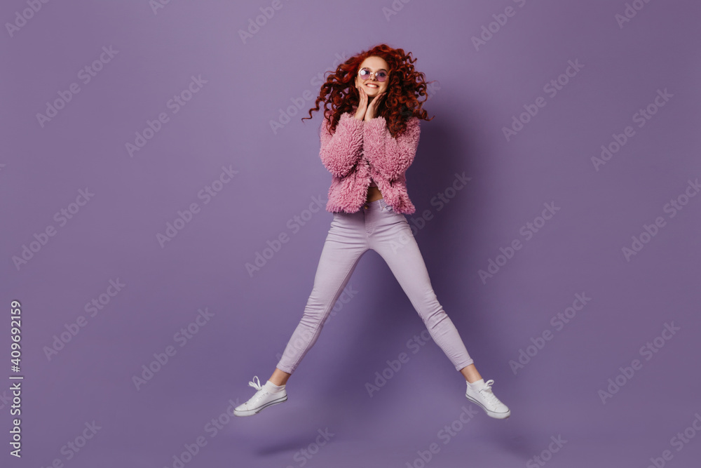 Smiling girl in round sunglasses jumps. Woman with red curls in bright outfit is having fun on purple background