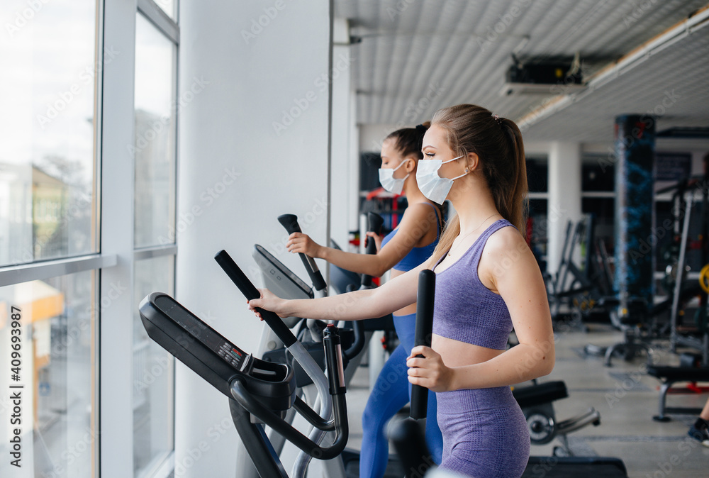 Fototapeta premium Two young beautiful girls exercise in the gym wearing masks during the pandemic. Social distancing in public places.