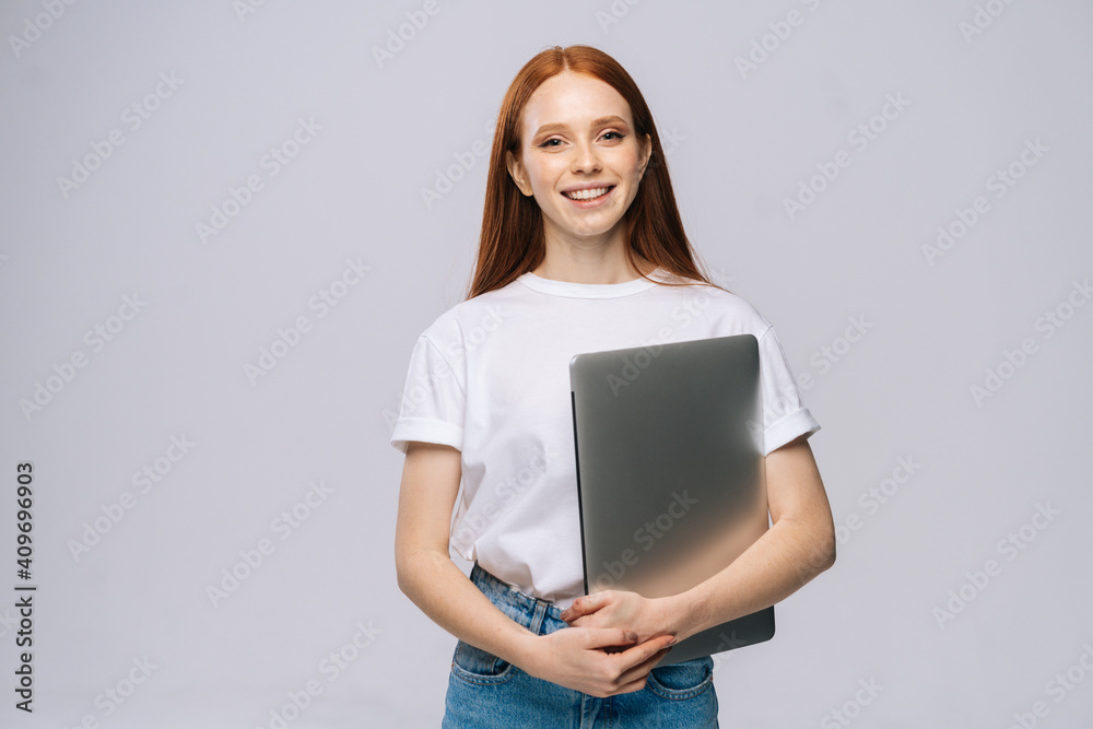 Charming young woman student holding laptop computer and looking at camera on isolated gray background. Pretty lady model with red hair emotionally showing facial expressions in studio, copy space. Charming young woman student holding laptop computer and looking at camera on isolated gray background. Pretty lady model with red hair emotionally showing facial expressions in studio, copy space.