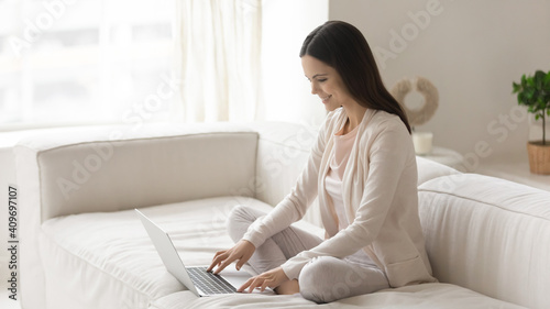 Smiling millennial Caucasian woman sit rest on sofa in living room text message on modern laptop. Happy young female relax on couch at home work online on computer. Technology, communication concept.