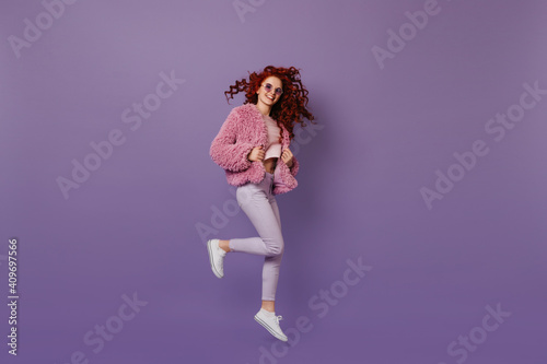 Charming woman in stylish light outfit and round glasses jumping. Girl with red hair is dancing, dressed in white jeans