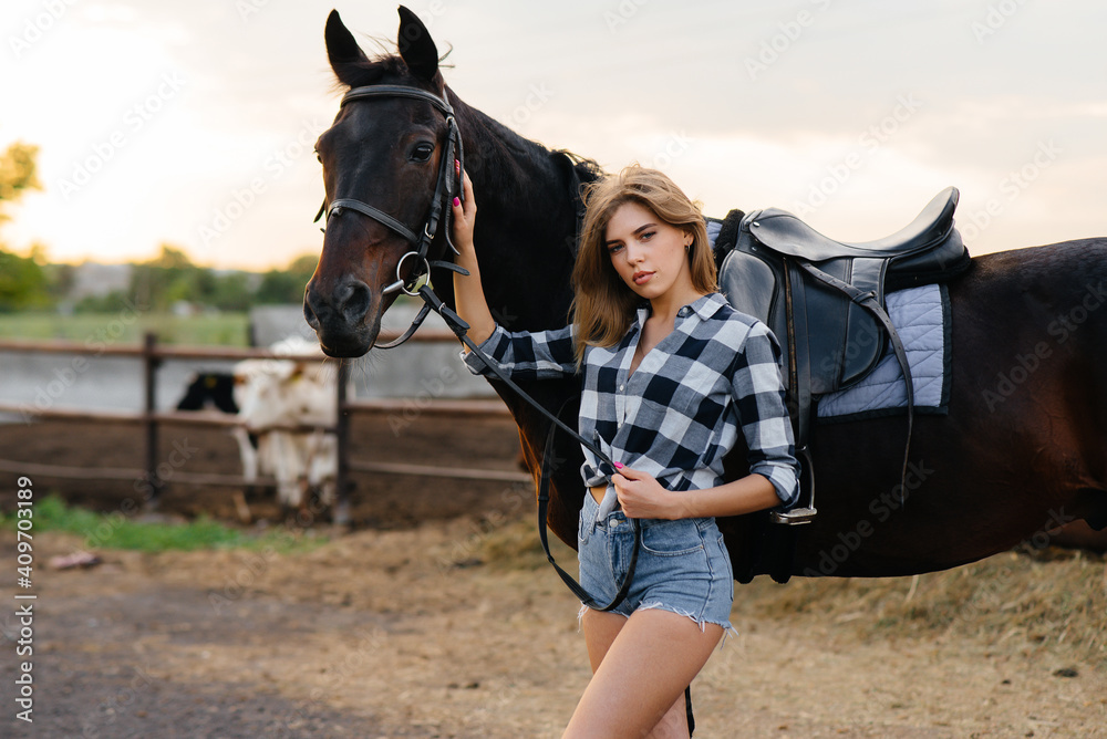 Obraz premium A young pretty girl rider poses near a thoroughbred stallion on a ranch. Horse riding, horse racing