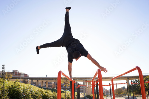 Man balancing with one hand, handstand on the bar, in a bar park in a city. Outdoor exercise. Concept of healthy living, sport, training, calisthenics.