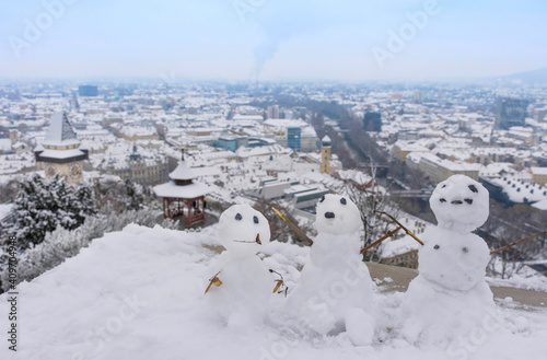 Three funny snowman and the famous clock tower on Schlossberg hill in the background  with snow  in Graz  Styria region  Austria  in winter day. Selective focus