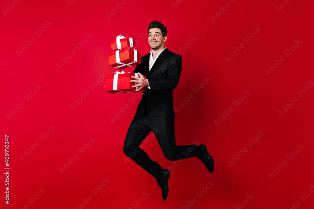 Full length shot of glad man jumping with valentine presents. Studio photo of brunette boy dancing on red background. Full length shot of glad man jumping with valentine presents. Studio photo of brunette boy dancing on red background.