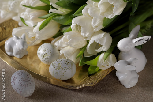 Decorative Easter eggs with ornaments  three figurines of Easter rabbits  made of ceramic  on a copper tray against a background of blurred white tulips