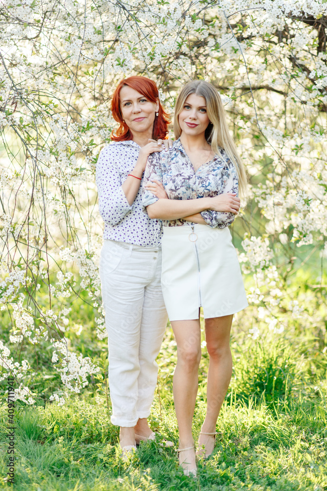 Fototapeta premium Two women, one young and the other older, in the spring in a beautiful garden with white flowers. Full-length photo