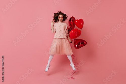 Studio shot of female model jumping with air balloons. Indoor photo of red-haired woman celebrating valentine's day.