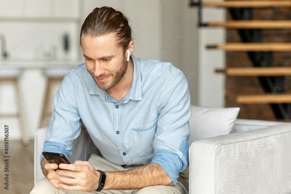 Stylish young bearded hipster guy is chilling at home, sitting on the comfy sofa in the modern living room, playing games, scrolling social media on the mobile phone, chatting with a friend, or dating Stylish young bearded hipster guy is chilling at home, sitting on the comfy sofa in the modern living room, playing games, scrolling social media on the mobile phone, chatting with a friend, or dating