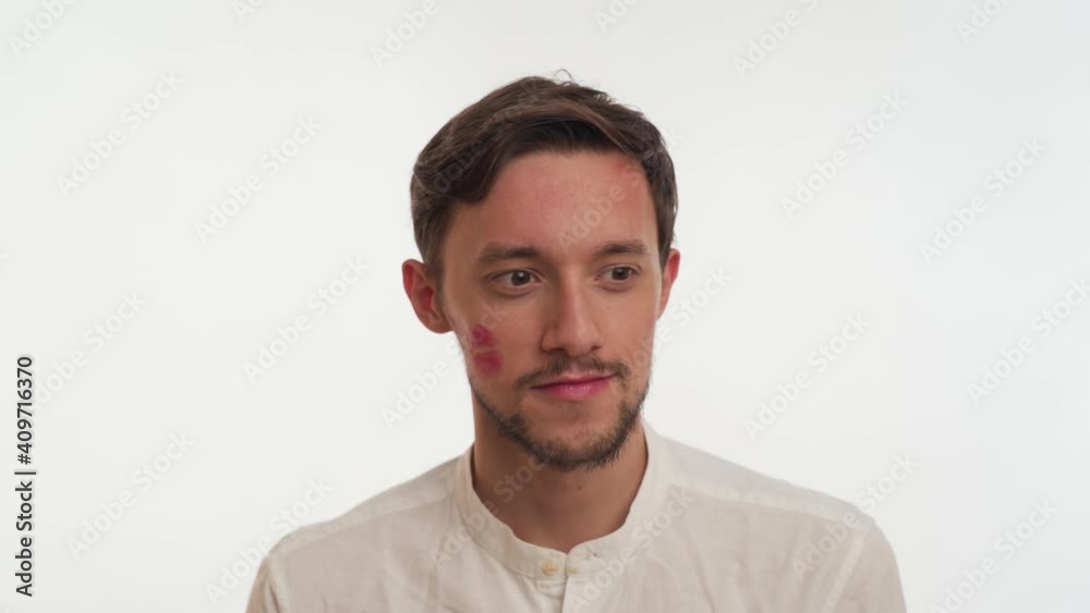 Scared handsome brunette Caucasian bearded man wear shirt, hold clear cotton pad, want to erase red lips stain of kiss from cheek by woman on white background close up. Concept of infidelity on wife. Scared handsome brunette Caucasian bearded man wear shirt, hold clear cotton pad, want to erase red lips stain of kiss from cheek by woman on white background close up. Concept of infidelity on wife.