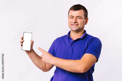 Caucasian man in blue t-shirt looks happy at the camera and shows his forefinger at the mocks up smartphone screen. Gray background. photo