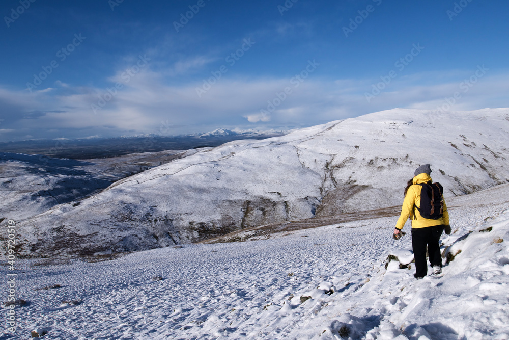 Active girl in a yellow jacket hiking in snow covered hills in Scotland on a beautiful sunny winter day
