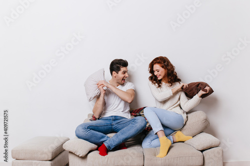 Indoor shot of couple enjoying pillow fight. Laughing family sitting on couch.
