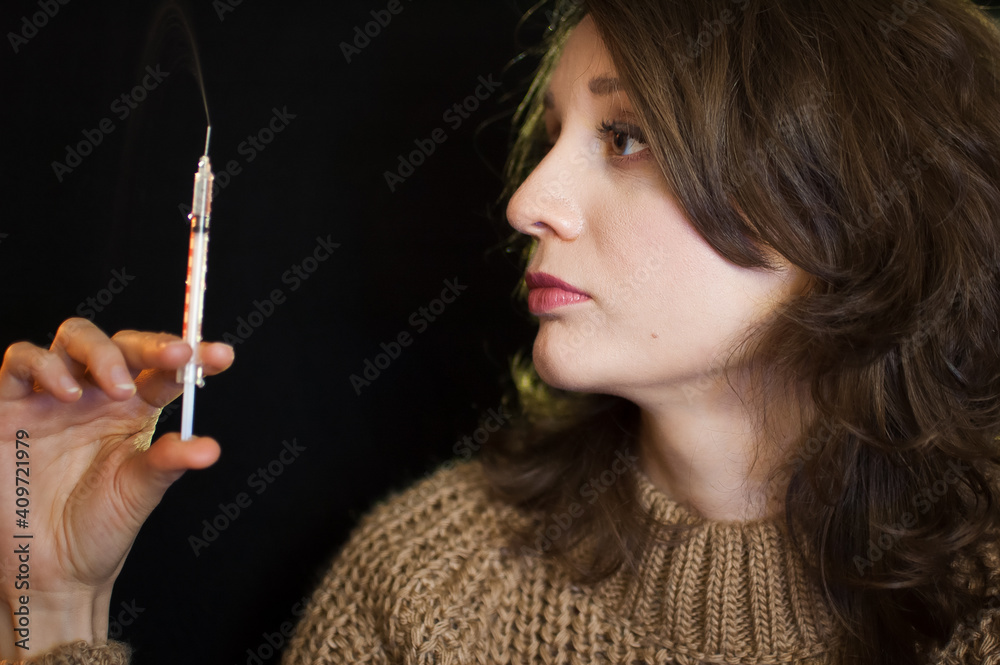 Obraz premium Vaccine concept. Female portrait of caucasian girl holding a syringe and looking at it on black background in studio.