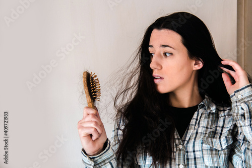 Mujer joven sujetando con la mano un cepillo con pelo caído mientras tiene una expresión de preocupación y mira frustrada. Concepto de caída del cabello