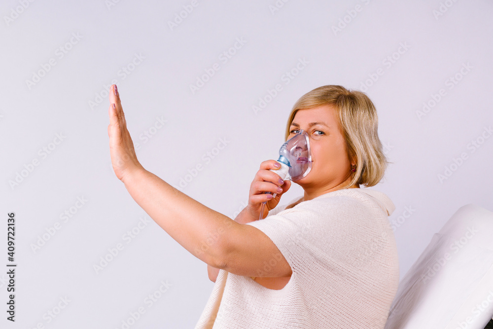The senior caucasian woman does inhalation with a nebulizer mask at home for coronavirus disease, showing stop sign on gray background.
