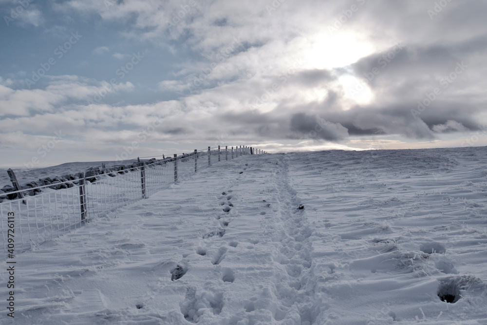 Path in snow next to a fence under cloudy sky with sunlight shining through